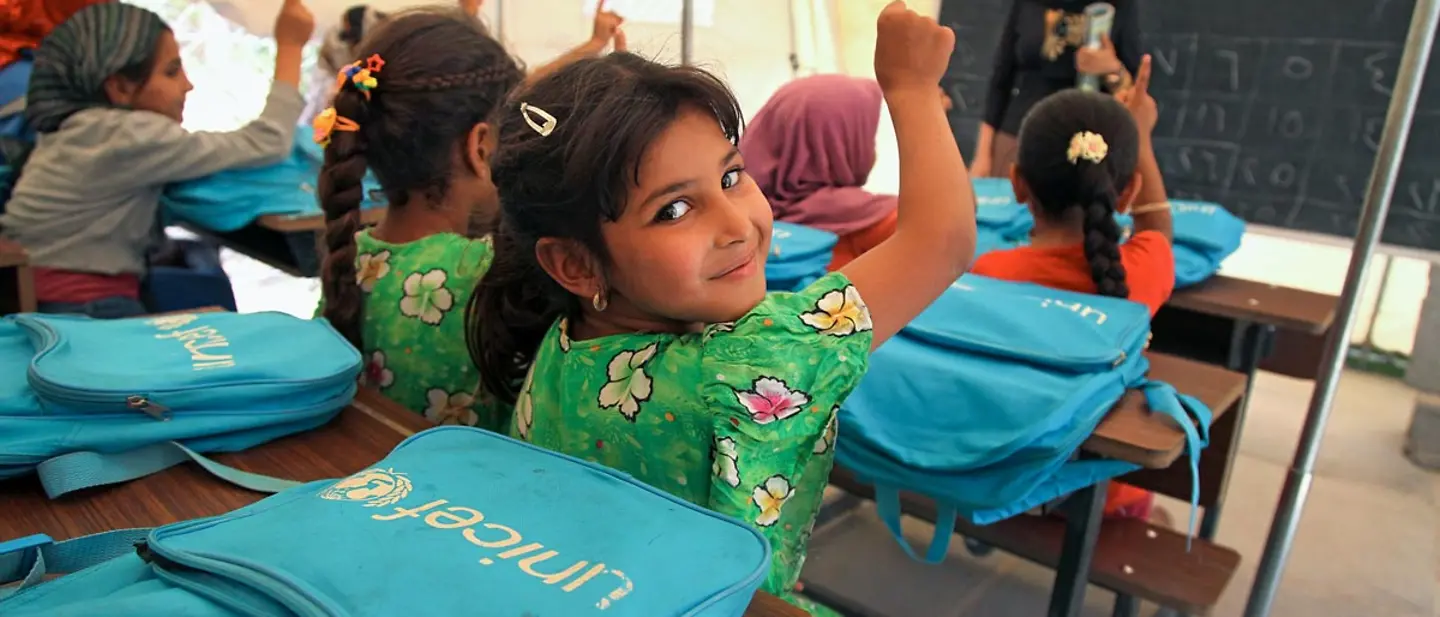 Kinder sitzen in einem Klassenzimmer und melden sich. Vor ihnen liegen Schulrucksäcke von UNICEF.
