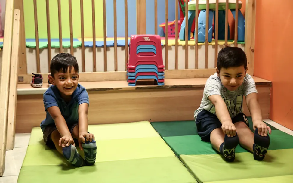 In dem Zentrum für Geflüchtete in Athen dürfen Kinder toben und finden außerdem neue Freunde. | © UNICEF/Karahalis Zwei Kinder machen zusammen Dehnübungen und freuen sich.