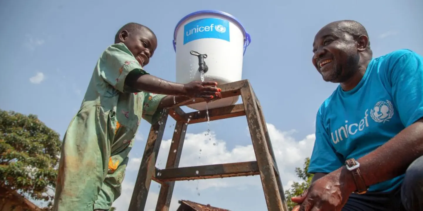 Ein Junge wäscht seine Hänge mit Wasser aus einem UNICEF Eimer neben einem UNICEF Mitarbeiter.