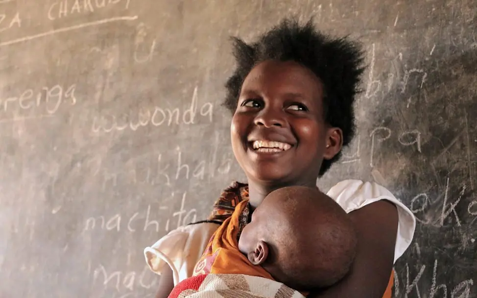 Amina hat an einem UNICEF-Alphabetisierungskurs teilgenommen. | © UNICEF/Chagara Schulen für Afrika: Eine junge afrikanische Frau steht mit ihrem Baby auf dem Arm in einem Klassenzimmer.