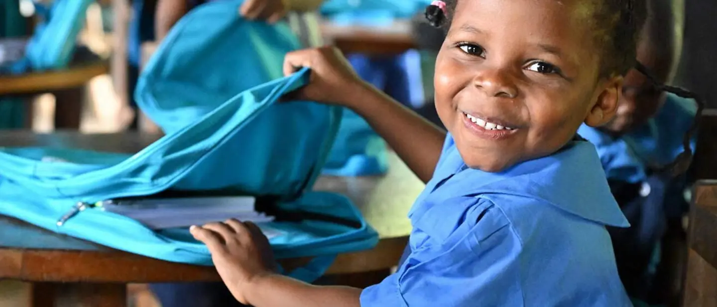Eine Grundschülerin in Kamerun öffnet lächelnd einen neuen Schulrucksack mit Schulmaterial.