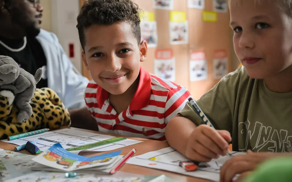 Weltkindertag einfach erklärt: Jungen sitzen mit Malsachen an einem Schultisch.   