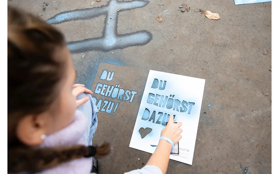 Ein Mädchen sprüht mit einer Schablone den Schriftzug "du gehörst dazu" am Boden mit Sprayfarbe auf.