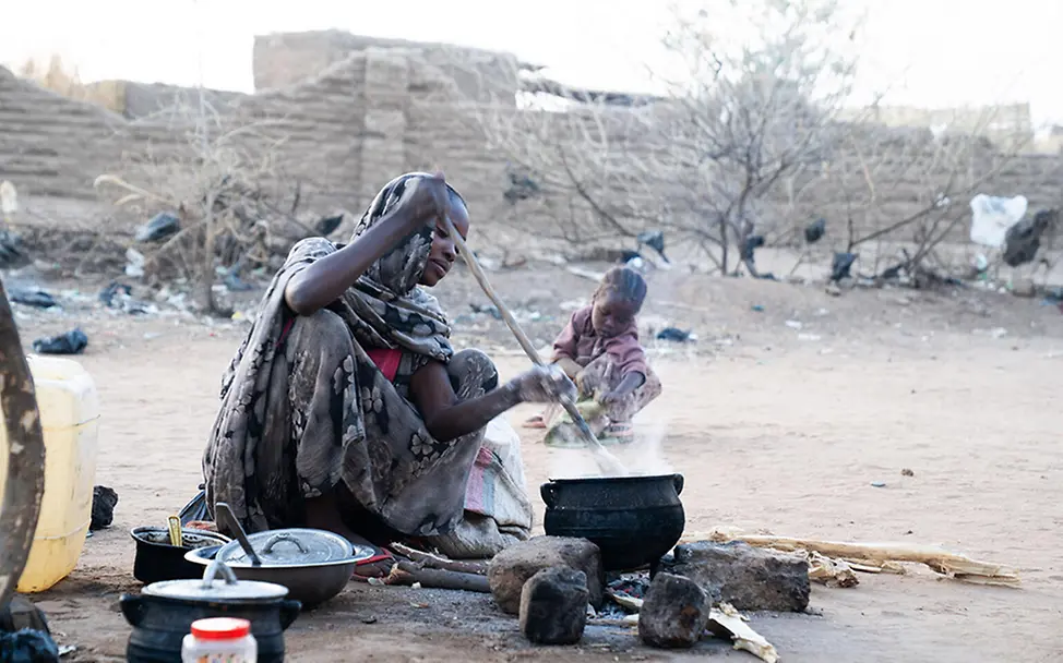 Eine Frau kocht im Sudan im Freien an einer Feuerstelle, während ein Kind in der Nähe im Staub sitzt.