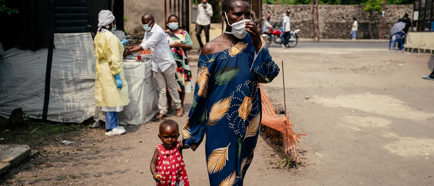 Eine Frau mit Atemschutzmaske geht mit ihrem Kind an der Hand an einer Gesundheitsstation im Kongo vorbei. | © UNICEF/AFP Ebola Kongo spenden: Eine Frau mit Atemschutzmaske geht mit ihrem Kind an der Hand an einer Gesundheitsstation im Kongo vorbei.