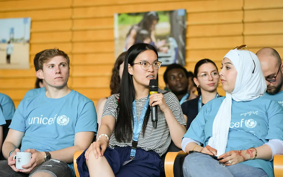 Engagierte der UNICEF-Hochschulgruppe Freiburg sitzen in einer großen Aula zusammen. | © UNICEF/Sachse-Grimm Engagierte der UNICEF-Hochschulgruppe Freiburg spricht beim Jahrestreffen in ein Mikrofon.