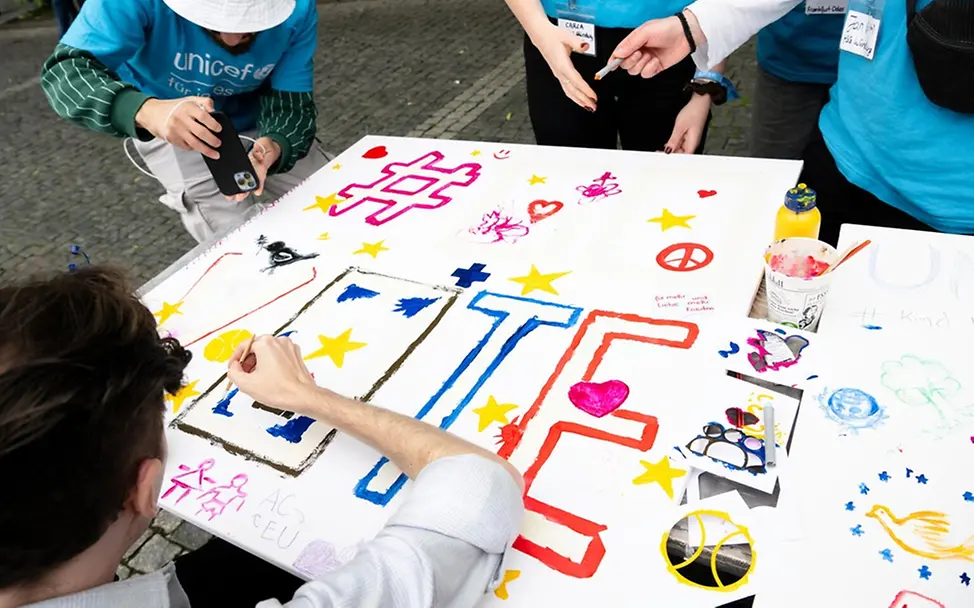 UNICEF-Hochschulgruppe aus Aachen beim Plakatgestalten. | © UNICEF/Etges Freiwilligenarbeit junge Erwachsene: Junge Menschen gestalten ein buntes Plakat, auf dem VOTE steht.