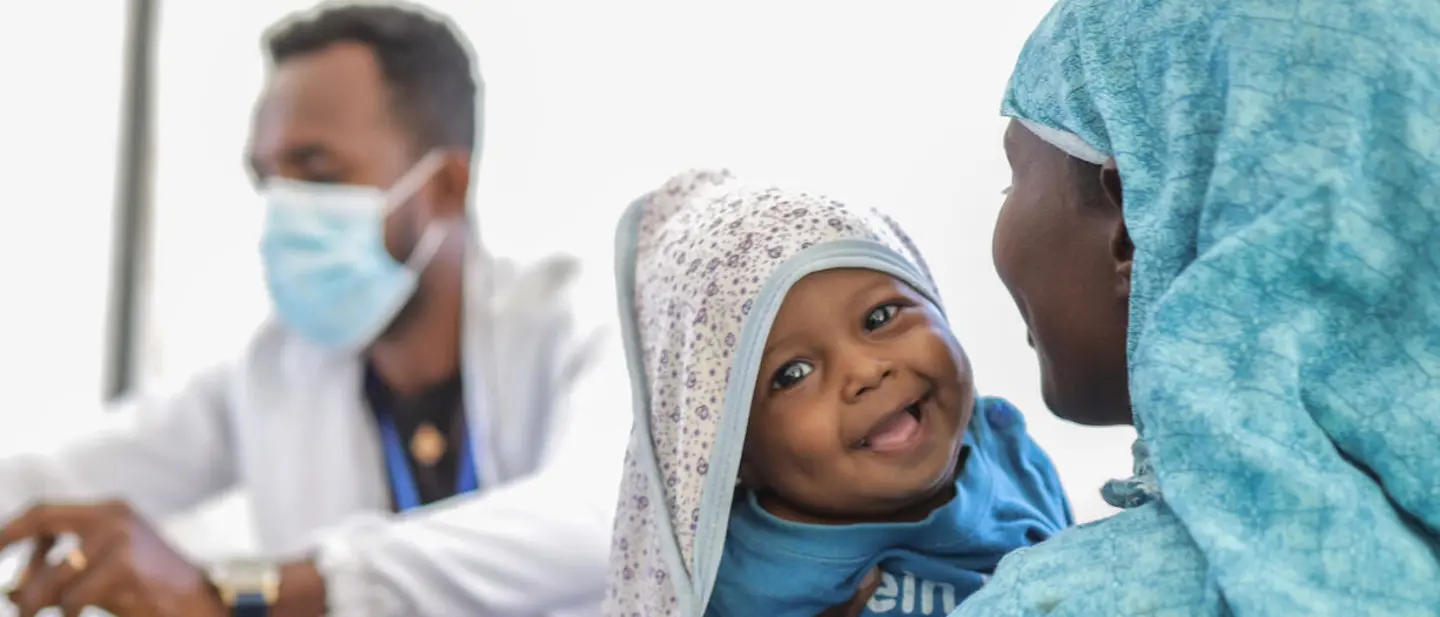 Geschäftsbericht 2025: Kind bei Untersuchung | © UNICEF/Tesfaye Geschäftsbericht 2025: In einem Behandlungsbereich hält eine Frau ein Kind im Arm; im Hintergrund sitzt ein Arzt mit Maske.
