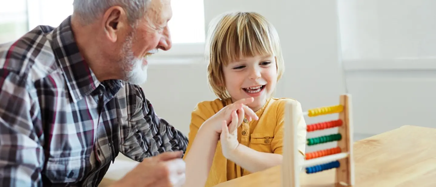 Mit einem Vermächtnis bedenken Sie gezielt bestimmte Personen, die eventuell keinen Erbanspruch haben. Vermächtnis oder Erbe: Ein Kind und ein älterer Mann sitzen lachend am Tisch und spielen.