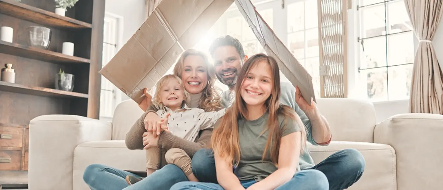 Der Pflichtteil schützt enterbte Angehörige, indem er ihnen einen Mindestanteil vom Erbe garantiert. Pflichtteil vom Erbe: Eine Familie sitzt gemeinsam auf dem Wohnzimmerboden unter einem vom Vater gehaltenen Dach aus Karton.