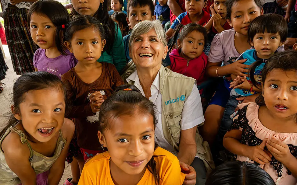 Guatemala: Eine Frau sitzt zwischen Kindern, die eng beieinander sitzen und teilweise lächeln oder aufmerksam schauen. | © UNICEF/Izquierdo Guatemala: Eine Frau sitzt zwischen Kindern, die eng beieinander sitzen und teilweise lächeln oder aufmerksam schauen.