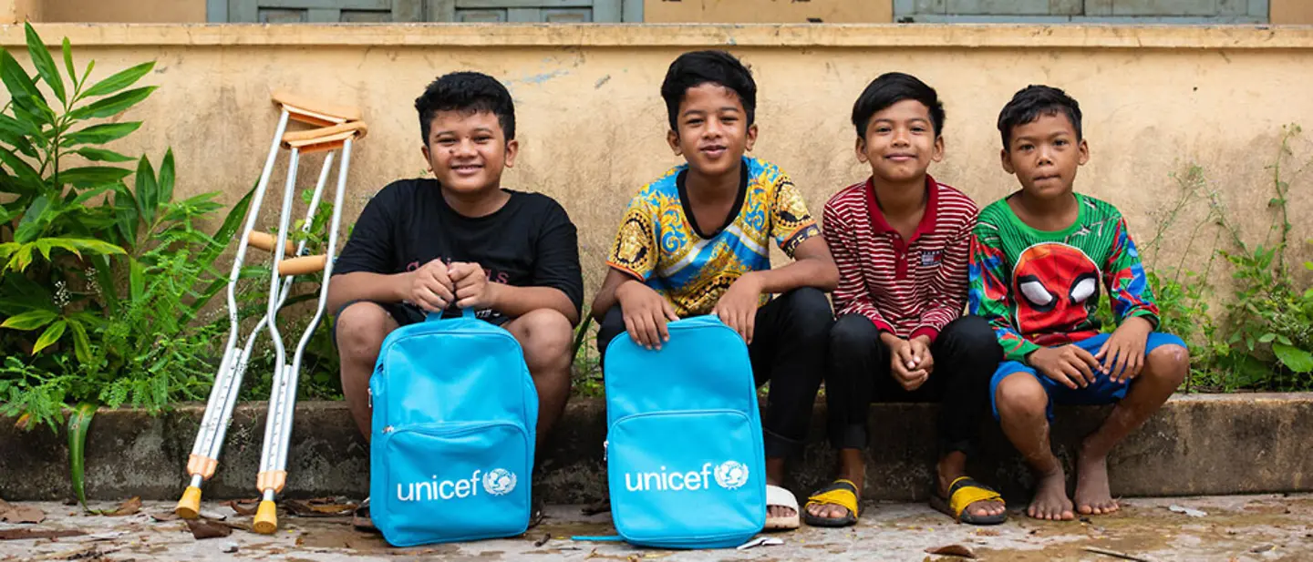 Kinder mit Schulrucksäcken in Kambodscha | © UNICEF/Wall Kambodscha: Vier Kinder sitzen vor einer Schulwand; zwei UNICEF-Taschen stehen davor, links liegen Krücken